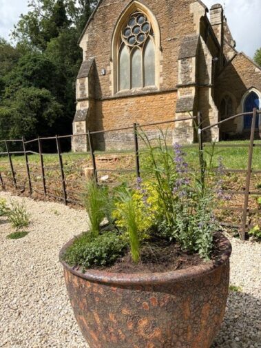 A large stone planter with green and purple plants sits in front of an old stone church, part of the Hidden Graves Project, with arched windows, a gravel path, and a metal fence. Trees are visible in the background.