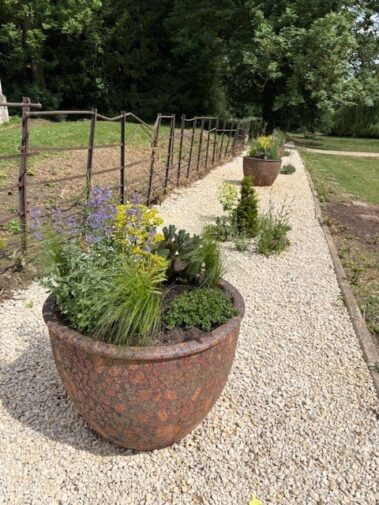 Large decorative planters filled with flowers and greenery line a gravel path next to a metal fence, marking the site of the Hidden Graves Project, with trees and grass visible in the background.