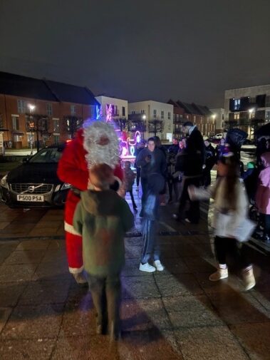 Santa Claus greets children in a festive outdoor setting at night, with people gathered around, colorful lights in the background, and parked cars and houses visible under a dark sky.