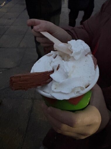 A person holds a green cup filled with vanilla ice cream, topped with a chocolate flake and a wooden spoon. The scene appears to be outdoors in low lighting.