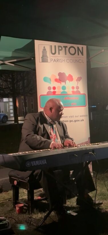 A man in a suit plays a Yamaha keyboard outdoors at night under a tent. Behind him is a sign for Upton Parish Council with colorful graphics and website information.