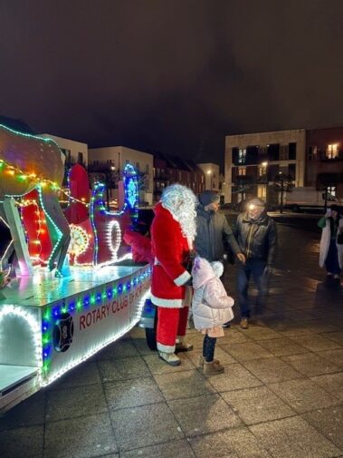 Santa Claus stands next to a child in winter clothes by a brightly lit, colorful float with reindeer decorations at night. People gather around, and buildings are visible in the background.