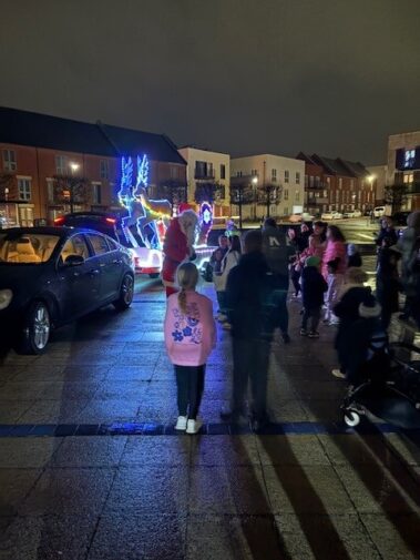 A group of people gather around a Santa Claus figure on a lit-up sleigh with reindeer at night in a residential area, with houses and parked cars in the background.