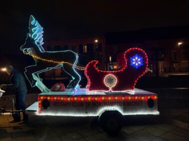 A festive parade float features a glowing reindeer and sleigh outlined with colorful lights, reading Rotary Club of Northampton Becket on the side, displayed outdoors at night.