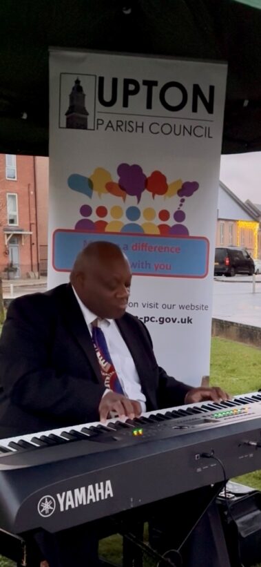 A man in a suit plays a Yamaha keyboard outdoors under a tent, with a Upton Parish Council banner in the background. Buildings and a car are visible behind him on a cloudy day.