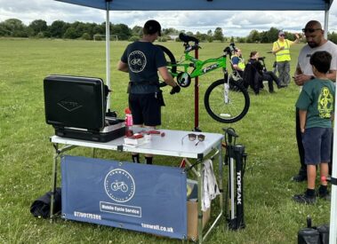 A mechanic repairs a green bicycle under a canopy at the Sports Fun Day with people nearby.