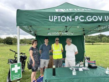 Four adults stand under a green Upton-PC.Gov.UK marquee at the Sports Fun Day beside first aid.